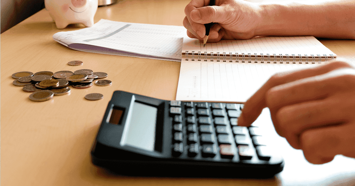 imagem de uma pessoa utilizando uma calculadora, um caderno  e varias moedas em cima da mesa.