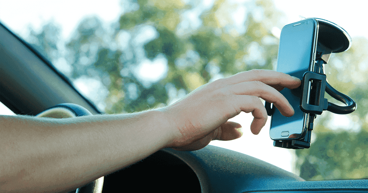 Imagem de um celular em um suporte no vidro do carro com uma pessoa utilizando o celular.